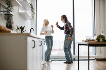 Caucasian old couple spends free time listening to music and dancing the twist in modern light kitchen. Seniors husband and wife having fun at the weekend in a cozy house.