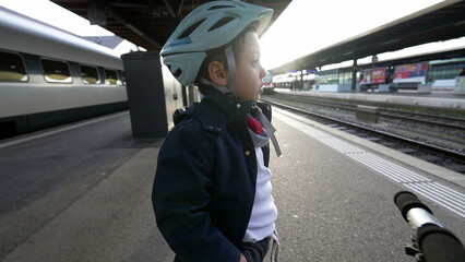 One pensive young boy standing at train platform wearing helmet staring blankly into the horizon...