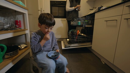 Young boy eating blueberries sitting in kitchen while mother puts away utensils and plates into...