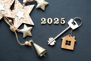 House key with keychain cottage on festive black background with stars, lights of garlands. New Year 2025 wooden letters, greeting card. Purchase, construction, relocation, mortgage, insurance