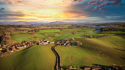 Aerial view of a picturesque village amidst green fields during sunset with a vibrant sky and a meandering river.