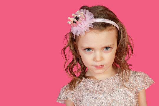 Portrait of serious little girl posing at pink background, sternly looking at camera. Cute emotional kid lady with blue eyes in pink dress, studio shot. Child image emotion concept. Copy ad text space