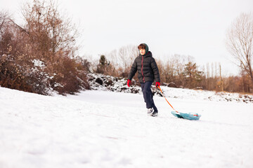 Teenager clad in a winter jacket vigorously pulls a bright teal sled across a snow covered landscape.Concept  winter sports, childhood, leisure activities during the cold season, and family fun.