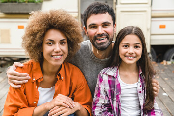 Cheerful mixed-race family of three parents and daughter looking at camera hugging together road...