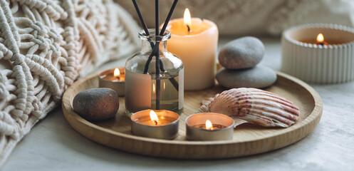 Fototapeta na wymiar Apartment natural aroma diffusor with sea breeze fragrance. Burning candles on bamboo tray, cozy home atmosphere. Relaxation, detention zone in the living or bedroom. Stones as decor. Banner