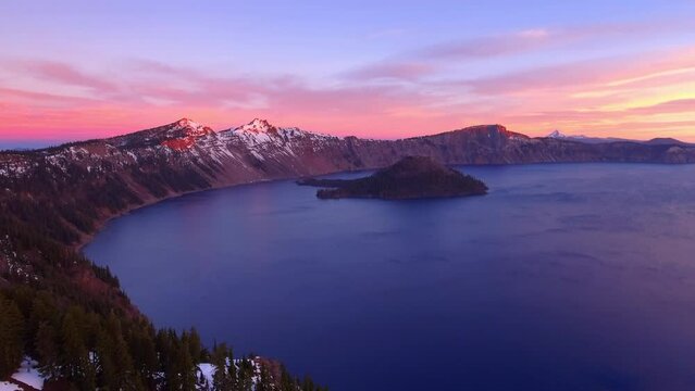 Aerial Scenic View Of Crater Lake National Park, Drone Flying Backwards Over Trees On Snowy Mountains During Sunset
