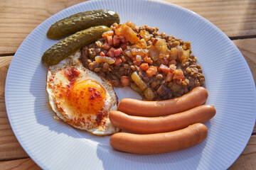 Close up picture on the white plate with traditional recipe from cuisine of Czech Republic. Lentils mash with roasted onion and pork sausages with sunny side up egg and sweet and sour pickled cucumber