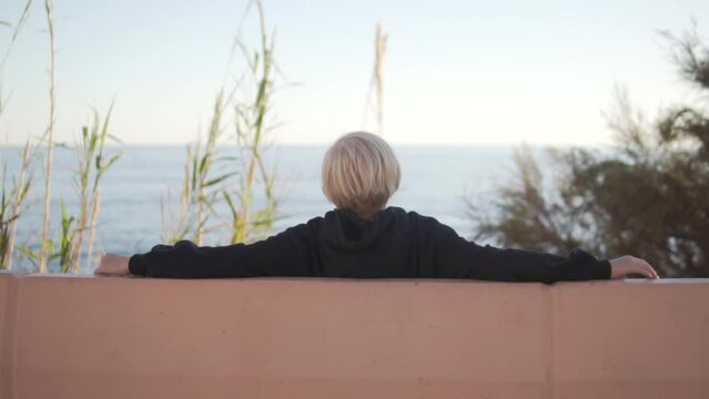 Blond boy sitting on a bench by the sea, view from the back, Spain, Alicante