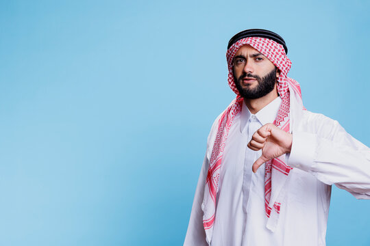Arab man wearing islamic clothes, showcasing serious expression and giving thumb down portrait. Serious muslim person showing disagreement gesture with finger and looking at camera