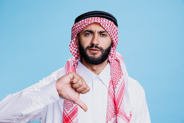 Muslim man dressed in traditional headdress showing thumb down negative gesture and looking at...