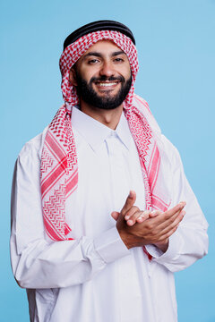 Muslim man wearing traditional clothes clapping hands and showcasing successful expression studio portrait. Cheerful arab dressed in islamic robe and headscarf applauding and looking at camera