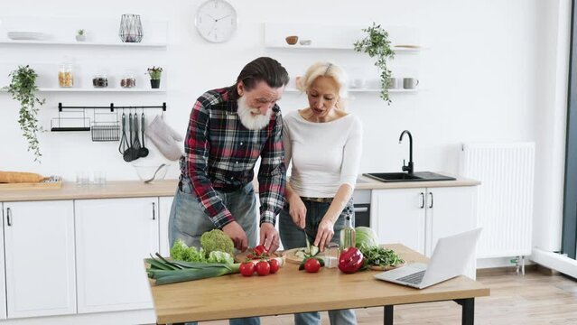 Caucasian old married man and wife prepare healthy salad, cutting thin slices of green vegetables and tomatoes with knife against background of spacious kitchen.
