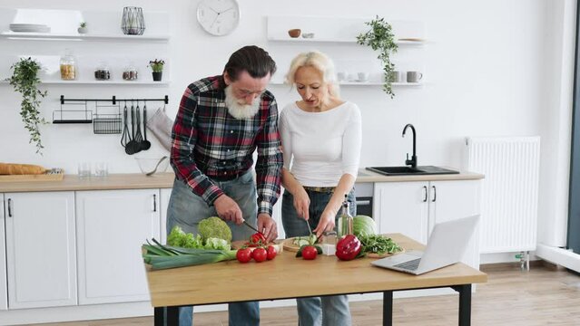 Caucasian old married man and wife prepare healthy salad, cutting thin slices of green vegetables and tomatoes with knife against background of spacious kitchen.