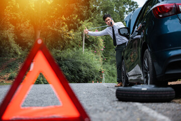 Asian businessman standing by broken down car, calling for help and showing thumbs up. Warning triangle behind car and man waiting for roadside assistance. Precautions and safety concept.