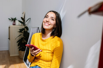 Smiling young beautiful woman with mobile phone relaxing at home. Joyful portrait of millennial female holding smart phone feeling happy.