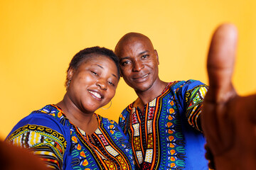 Smiling black man and woman wearing ethnic clothes taking selfie on smartphone and looking at front camera. Cheerful couple holding mobile phone and making photo pov portrait - obrazy, fototapety, plakaty
