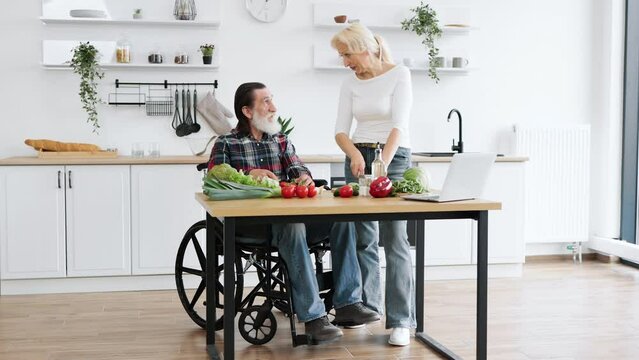 Happy old married man in wheelchair and wife prepare healthy salad, cutting thin slices of greenery vegetables and tomatoes with knife against background of spacious kitchen.