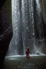 A young, slender woman in a pink swimsuit swims in an incredibly beautiful mystical waterfall in a cliff on the popular island of Bali.