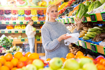 Obraz premium Interested woman shopping for leafy vegetables in supermarket, choosing fresh green romaine lettuce on shelves..