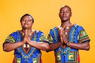 Fototapeta na wymiar Black man and woman with closed eyes standing with folded palms, praying and asking for blessing. African american couple wearing ethnic clothes meditating and relaxing together