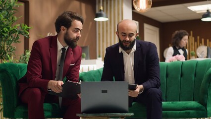 People in telework session online undertaking business meetings while seated at hotel entrance lounge. On a video conference, supervisory team interacts with front desk and remote staff members.