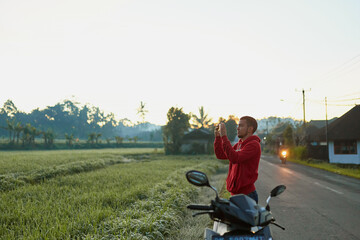 A young man stands on the side of the road near a rice paddy and takes a picture with his phone of Mount Agung on the popular tourist island of Bali.