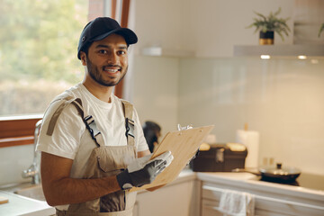Portrait of young smiling repairman standing with clipboard in kitchen at home - obrazy, fototapety, plakaty