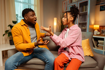 Young emotional African American couple, man and woman quarreling sitting in room at home conflict