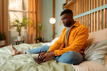 Attractive African American man, professional programmer sitting on bed, using laptop, at home