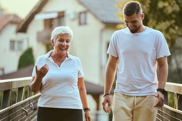 A handsome man and an older woman share a serene walk in nature, crossing a beautiful bridge against the backdrop of a stunning sunset, embodying the concept of a healthy and vibrant intergenerational