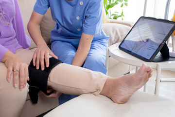 Caregiver or nurse helping check tendon and arthritis of knee or leg for diagnostic and rehabilitation while explaining senior woman with tablet living room at home, caretaker or physiotherapist.