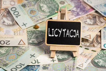 Black writing board on a wooden frame with the inscription "licytacja", Polish zloty PLN banknotes scattered in the background (selective focus) translation: auction