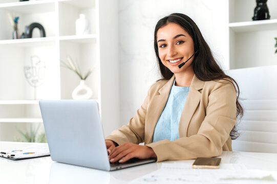 Photo of a confident positive woman, call center operator with headset, working on support hotline in a modern office with a laptop, looking at the camera, smiling friendly. Customer support service