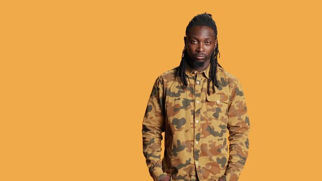 Portrait of man with braids smiling in front of camera, wearing cool camo clothes and feeling happy in studio. African american person with trendy hair posing over orange background, relaxed style.