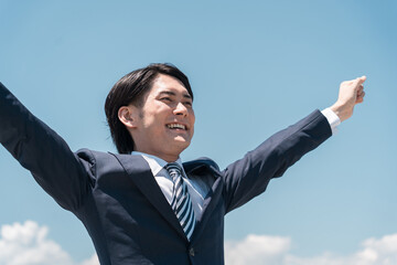 青空の中で喜ぶ若いアジア人ビジネスマン（転職・勝利・就職・脱サラ・チャレンジ・頑張る・達成）
