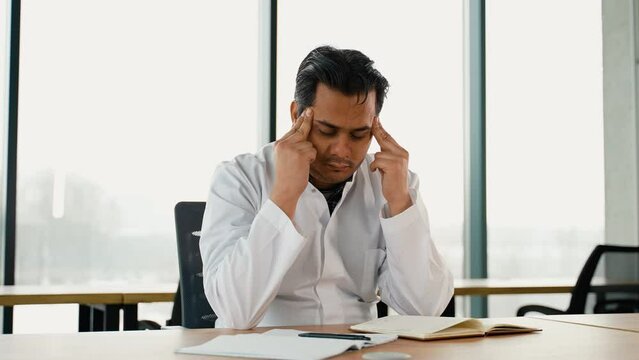 Stressed frustrated Indian doctor with headache sitting alone at work desk in hospital. Sad young gp feeling desperate and guilty for medical failure