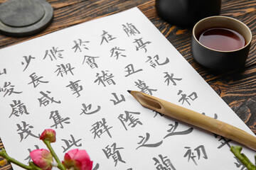 Paper sheet with Asian hieroglyphs, nib pen and cup of tea on wooden table, closeup. International...