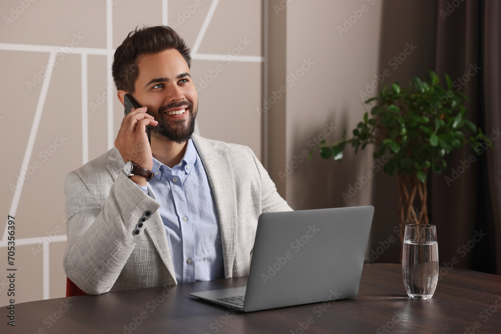 Canvas Prints Happy man using modern laptop while talking on smartphone at table in office