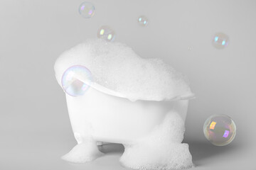 Small bathtub with foam and soap bubbles on grey background - obrazy, fototapety, plakaty