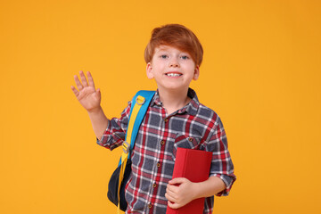 Happy schoolboy with backpack and book waving hello on orange background