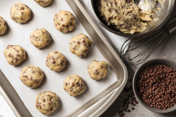 Baking pan with raw chocolate chip cookie balls, ingredients and whisk on table, flat lay