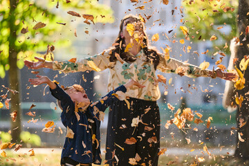 A modern woman joyfully plays with her son in the park, tossing leaves on a beautiful autumn day, capturing the essence of family life and the warmth of mother-son bonding in the midst of the fall