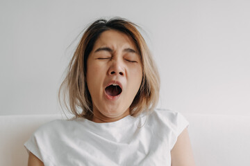 Sleepy face Asian woman yawning and rubbing her eyes in white t-shirt sits on the sofa.