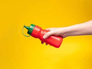 Hand holds red ketchup bottle with green lid against cheerful yellow background, ideal for food concepts. 
