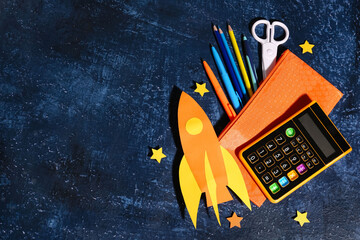 Orange paper rocket with calculator, pencil case and scissors on dark grunge background
