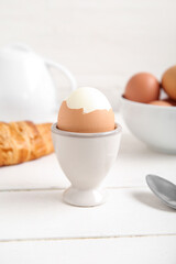 Holder with boiled chicken egg, teapot and croissant on white wooden background