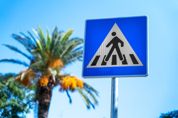 Close-up of road sign square blue pedestrian crossing pointer on background of tropical trees, palm trees and clear sky on sunny day Road intersection sign in a public tourist place, beach Resort sign