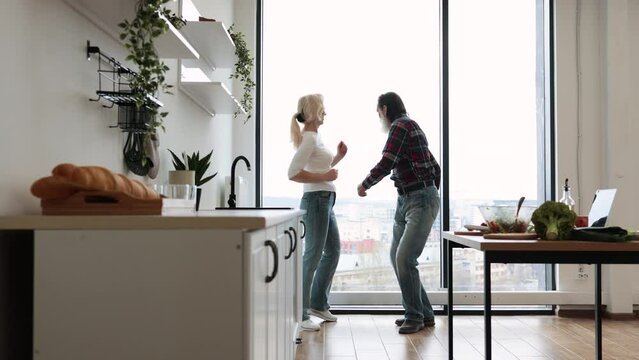 Seniors husband and wife having fun at the weekend in a cozy house. Caucasian old couple spends free time listening to music and dancing twist in modern light kitchen.