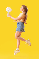 Young woman with electric fan jumping on yellow background