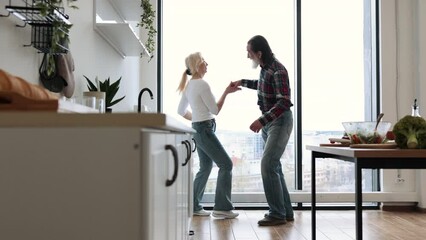 Seniors husband and wife having fun at the weekend in a cozy house. Caucasian old couple spends free time listening to music and dancing twist in modern light kitchen.